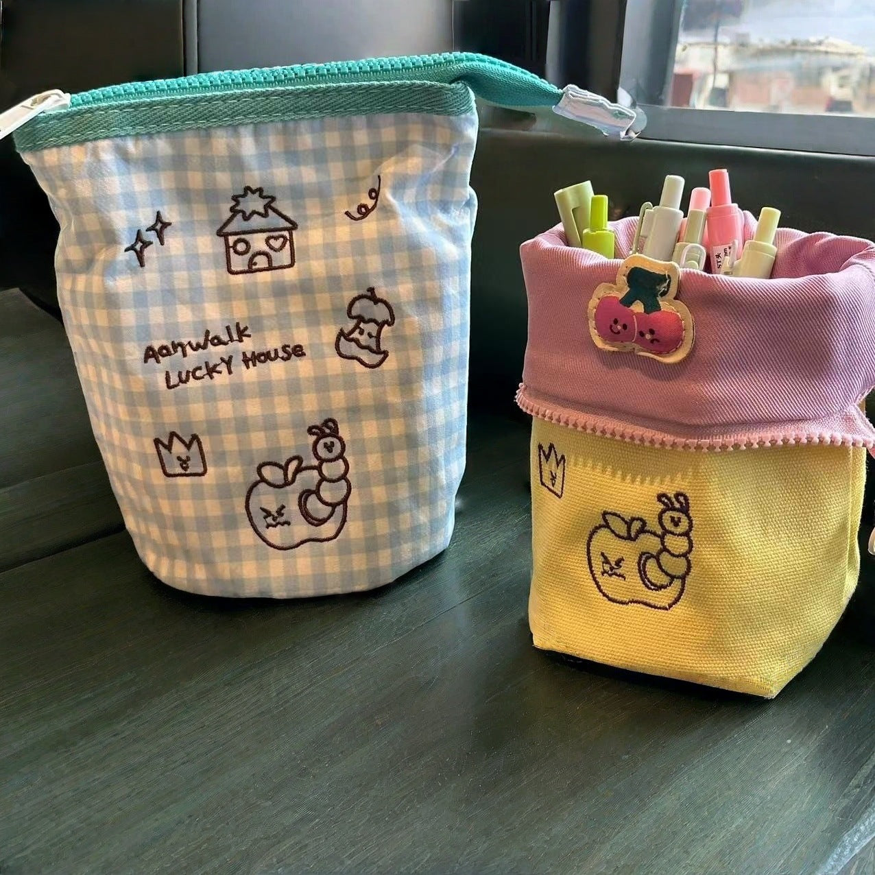 Simple Vertical Flip large capacity pencil cases with cute embroidered designs and pastel colors on a dark surface near a window